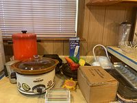 Photo showing various kitchen items: white Crock-Pot slow cooker, orange container, boxed carving knife, George Foreman grill, potato baking dish, coffee brewer without cord, box, slicer, and decorative ceramic fruits.