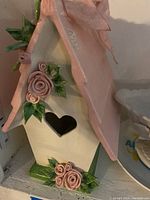 Close-up of pink and green porcelain birdhouse showing heart-shaped entrance and rose decorations on roof and base