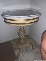 Full side view of the round table showing white marble top with slight veining and light wood base carved into pineapple shape with a round pedestal and square platform base with legs.