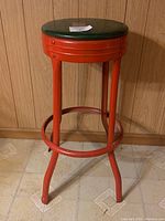 Front angle view of the red metal bar stool with green vinyl seat, showing overall form and structure.