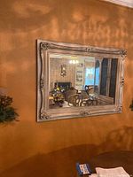 Front view of large vintage French Provincial wall mirror hanging on an orange wall reflecting living room with furniture and ceiling fan.