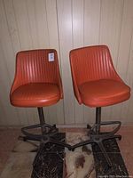 Two orange vinyl Mid-Century Modern swivel barstools with vertical stitched lines in the seats and backs, set against a paneled wall on marble flooring.