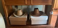 Two western-style hats resting on white cardboard boxes under a wooden table, one light-colored and one dark brown.