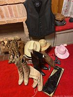 Lot of western wear including 3 vests, several cowboy hats, and multiple pairs of ladies boots on a red carpet in front of brick fireplace.