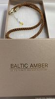 Gold-colored twisted cord necklace displayed coiled inside a Baltic Amber branded box with cushioning material.