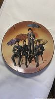 Front view of the 8.5-inch Beatles collector plate with all four members seated/standing holding umbrellas