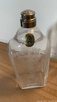 Front view of the German crystal glass flask showing the etched decorative floral and scroll pattern, metal cap, and brand label near neck.