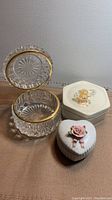 Image showing three trinket boxes: a clear crystal box with attached lid and gold trim, a hexagonal ceramic box with floral lid, and a white ceramic heart-shaped box with pink rose decoration.