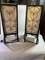 Two black iron work lamps with scroll designs and natural mica shades, standing side by side on white surface.