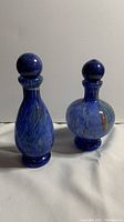 Two Murano style blue art glass bottles side by side on a white surface with visible colors and stoppers