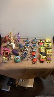 Wide shot showing the full assortment of 26 plastic toys including SpongeBob, A Bug's Life characters, and Lilo & Stitch bobbleheads arranged on a table.