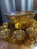 Front view of punch bowl surrounded by nine matching cups showing marigold iridescent finish and embossed grape motif detailing.