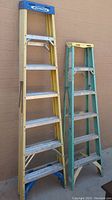 Two Werner fiberglass step ladders leaning against wall, one yellow 7 ft, one green 6 ft, showing wear and use marks.