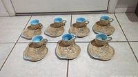Seven tea cups on matching saucers, arranged on a tiled floor showing the set as a whole.