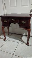 Front view of wooden vanity/desk with three drawers and brass handles, showing curved legs and scalloped apron