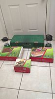 Full view of the lot showing two boxed Coleman Quickbed queen air mattresses, two green Coleman lanterns, and a green metal portable camping stove with latch.