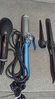 Photo showing all three styling tools: black paddle brush straightener, blue clamp curling iron, and black curling wand attachment, with cords coiled.