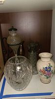 Photo of Polonia lead crystal vase, two vintage oil lamps, copper/brass pitcher, and ceramic vase on shelf