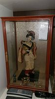 Full view of geisha doll inside wooden and glass display case with textured gold background.