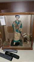 Front and side view of the geisha doll standing inside a wooden and glass display case with a miniature setting including small figurines and a tiny structure. Remote controls placed near the case to indicate scale.