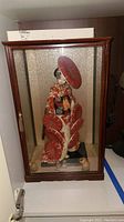 Full front view of the Geisha doll in the wooden glass case showing multiple red parasols and decorative figures inside.