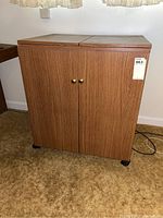 Front view of a wooden sewing table cabinet with two front doors and brass-colored knobs, placed on beige carpet.