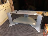 Metal TV stand with silver metal frame and one intact frosted glass shelf in lower center. Top glass shelf is missing revealing metal supports.