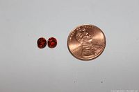 Two small round cut garnet gemstones shown beside a penny for size reference. Gems appear deep red-orange and are faceted.