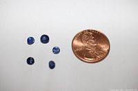 Top view of 5 loose faceted blue sapphires next to a penny for size reference. Stones vary in size with deep blue color and round cut facets.