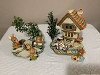 Overall lot view showing two separate ceramic bases with bunny figurines and greenery; one base features a large house and two bunnies, the other base shows three bunnies with a water feature.