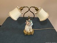 Full front view of the antique porcelain and brass dual-shade lamp on a table, showing the dual heart-shaped brass support and fabric shades.