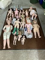 Entire collection of twenty-one porcelain dolls displayed on a brown surface, including dolls of varying sizes and states of dress, doll stands, and some accessories scattered around.