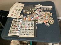 Photo showing a large stamp album open with many used stamps affixed on pages. Numerous loose stamps and small paper pieces spread on the table around the album.