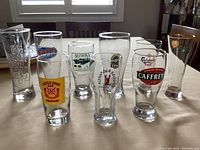 Front row and back row view of nine assorted branded beer glasses on a wooden table with window light.