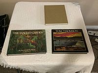 Photo showing three large hardcover art books placed on a white textured surface: 'The Tangled Garden' by J.E.H. MacDonald, 'Tom Thomson: The Silence and The Storm', and a plain tan hardcover book (likely Emily Carr).