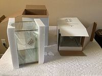 Two Bowring branded boxes with packaging foam on a textured white surface holding two glass hurricane candle holders, one with silver trim and one with gold trim, both unboxed and protected.