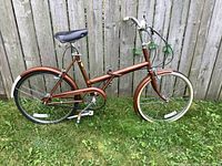 Side view of the entire vintage Raleigh fold-up bicycle showing brown frame, wheels with reflectors, black saddle, and cream handlebar grips.