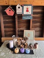 Photo shows three wooden birdhouses mounted on wooden boards, wooden votive candle holders, various candles, and framed decorative needlework pieces on a table.