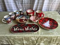Full set showing combination of Santa themed plates, red and white striped cups and bowls, red mugs, and holiday serving platters on a festive tablecloth.