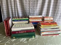 Photo of entire lot showing three main stacks of cook books including a boxed set, thick hardcover cook books and softcover magazines and books.