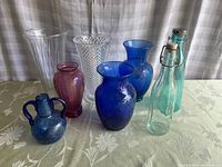 Full view of all 8 pieces showing variety of colors and shapes including cranberry, blue, clear glass vases and bottles with flip-top caps.