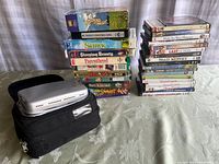 Portable DVD player and carrying case on table with stacks of DVDs and VHS tapes.