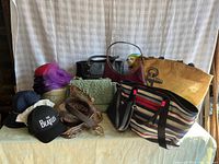 Overview of assorted handbags, totes, caps, hats and belt arranged on table against curtain background.