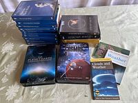 Photo showing the complete lot including DVDs stacked vertically and horizontally along with the two weather topic books lying flat on a tablecloth.