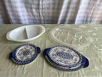 Four serving platters on a table: two blue Fine-King Ware ceramic platters with floral pattern, one white divided ceramic platter, and one clear Pyrex glass oval platter with section divisions.
