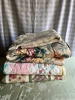 Stack of four folded textiles including one beige throw blanket on top and three quilts with floral and patchwork designs below.