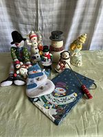 Wide shot of assorted snowman figurines including wooden, fabric, and plastic materials, plus the Russ brand snowman wall art on table against draped backdrop.