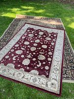 Two stacked polypropylene rugs on grass; the larger one has dark intricate floral design and the smaller one on top has burgundy center and beige border with floral motifs.