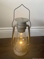 Photo of a battery-operated plastic lantern with a clear globe and beige colored body. The lantern is lit and has a handle for carrying or hanging.