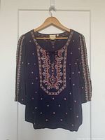 Front view of Anthropologie embroidered deep purple top hanging on wooden hanger against white door, showing detailed embroidery and beading on front panel and sleeves.